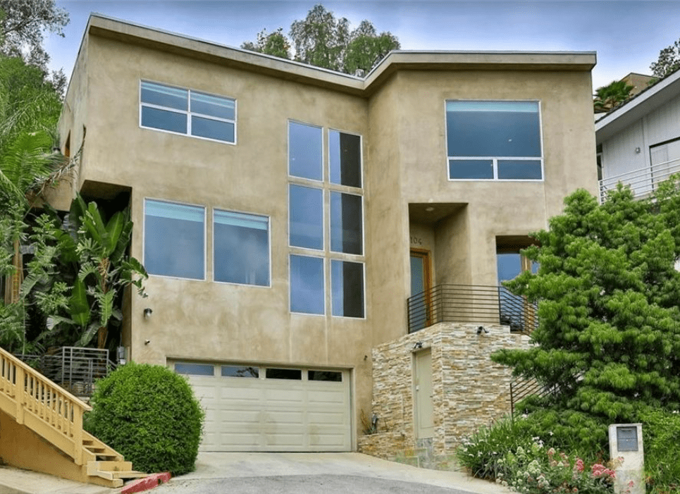 A luxurious three-story modern home with beige stucco siding, large picture windows, and a lower-level garage. There are stairs leading up to the entry door on the second floor. The house is surrounded with greenery.